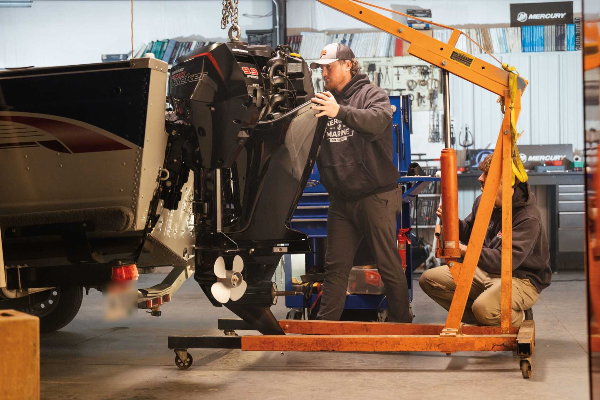 Two technicians mounting a Mercury outboard engine on a boat in a workshop